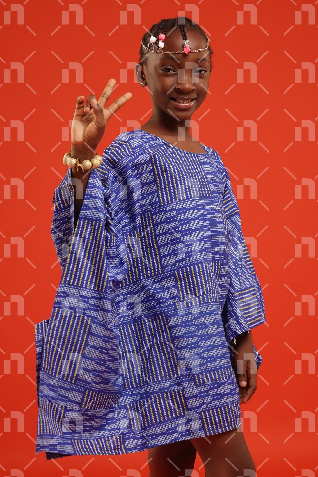 fillette-africaine-moderne-vetue-dune-robe-de-couleur-bleu-posant-legerement-de-profil-avec-deux-doigts-de-main-droite-leves-et-un-grand-sourire-pour-une-photo-studio
