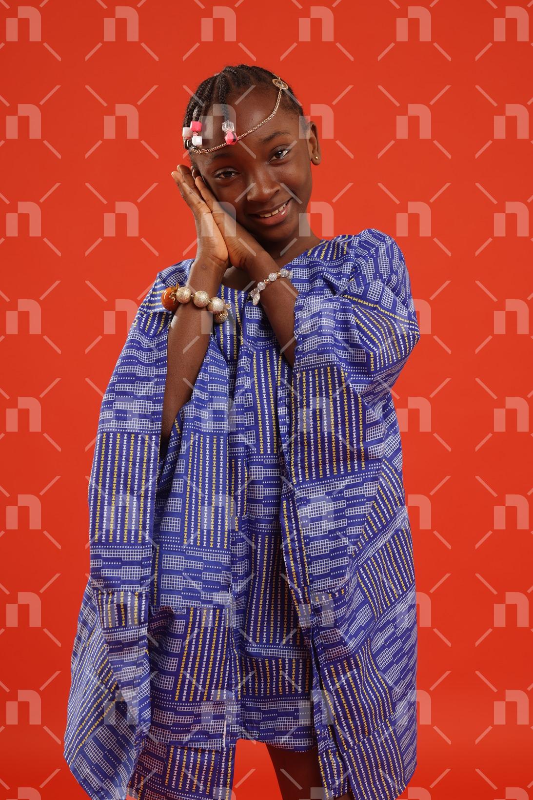 fillette-africaine-moderne-vetue-dune-robe-de-couleur-bleu-dans-une-posture-la-tete-posee-sur-les-deux-mains-jointes-sous-la-joue-avec-un-grand-sourire-pour-une-photo-studio