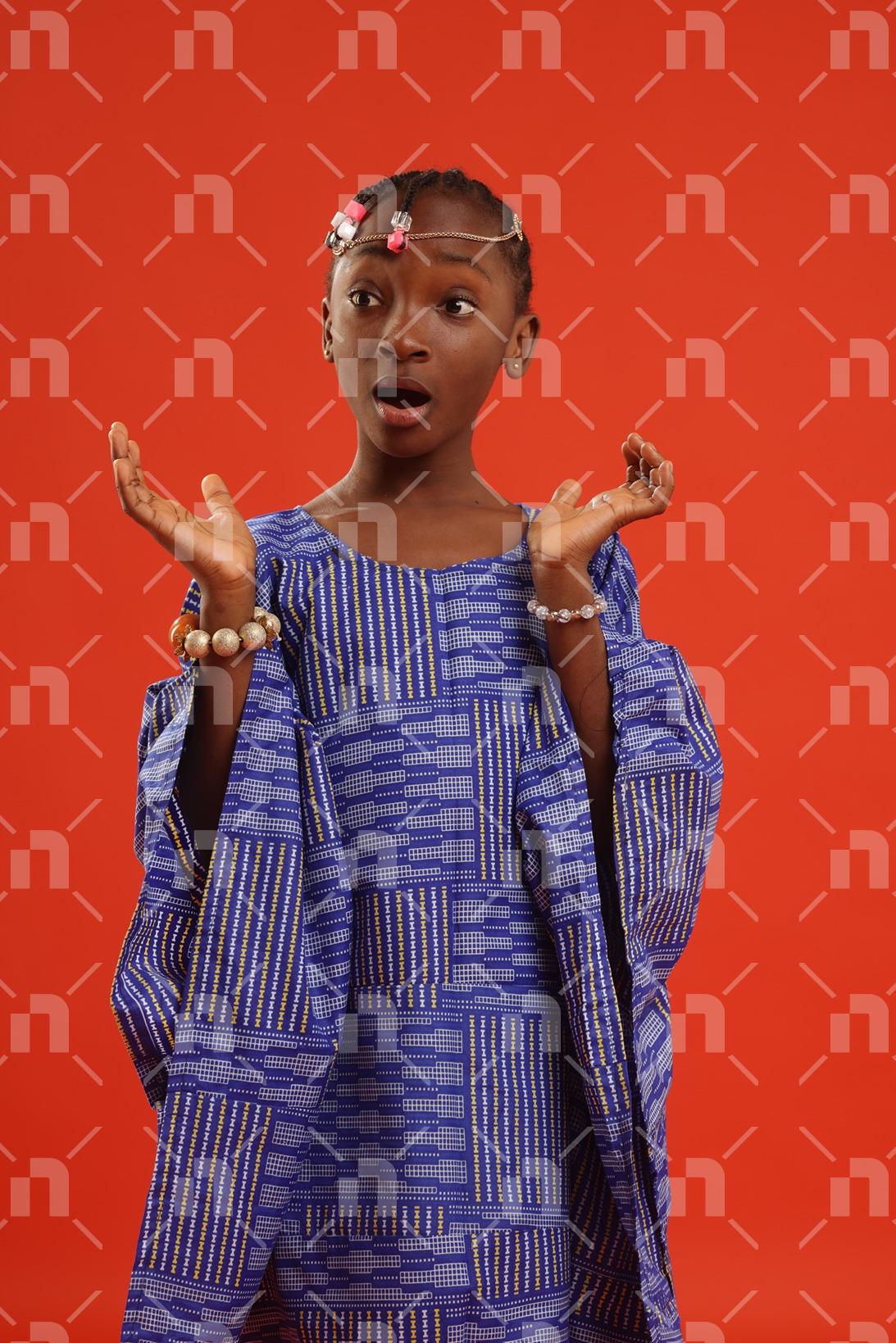 fillette-africaine-moderne-vetue-dune-robe-de-couleur-bleu-executant-un-geste-dinnocence-avec-les-yeux-fixes-sur-la-droite-et-une-bouche-ouverte-pour-une-photo-studio