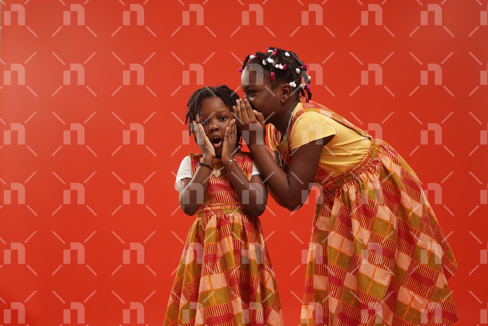 deux-filles-dont-lage-varie-entre-5-et-10ans-portant-des-robes-faites-en-pagne-traditionnel-de-couleur-orange-dont-lune-chuchote-a-loreille-de-la-plus-petite-avec-le-sourire