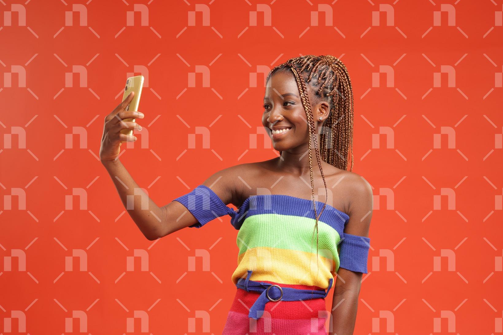 jeune-fille-africaine-moderne-vetue-dune-robe-coloree-prenant-un-selfie-a-laide-de-son-telephone-sur-un-fond-orange