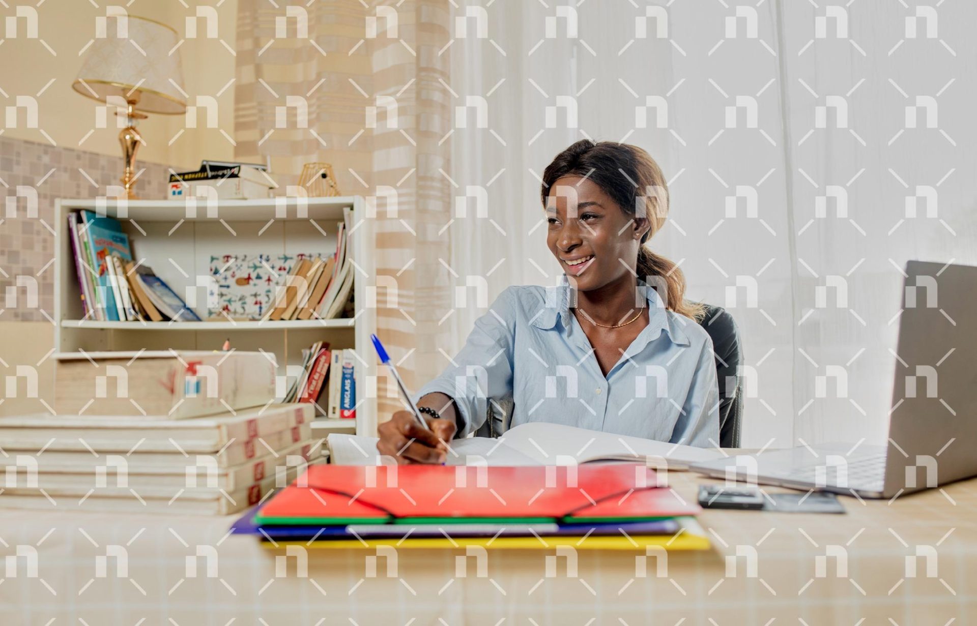 la-tete-tournee-vers-la-droite-une-jeune-femme-africaine-souriante-ecrivant-dans-un-grand-cahier-a-son-bureau-avec-un-ordinateur-portable-a-cote