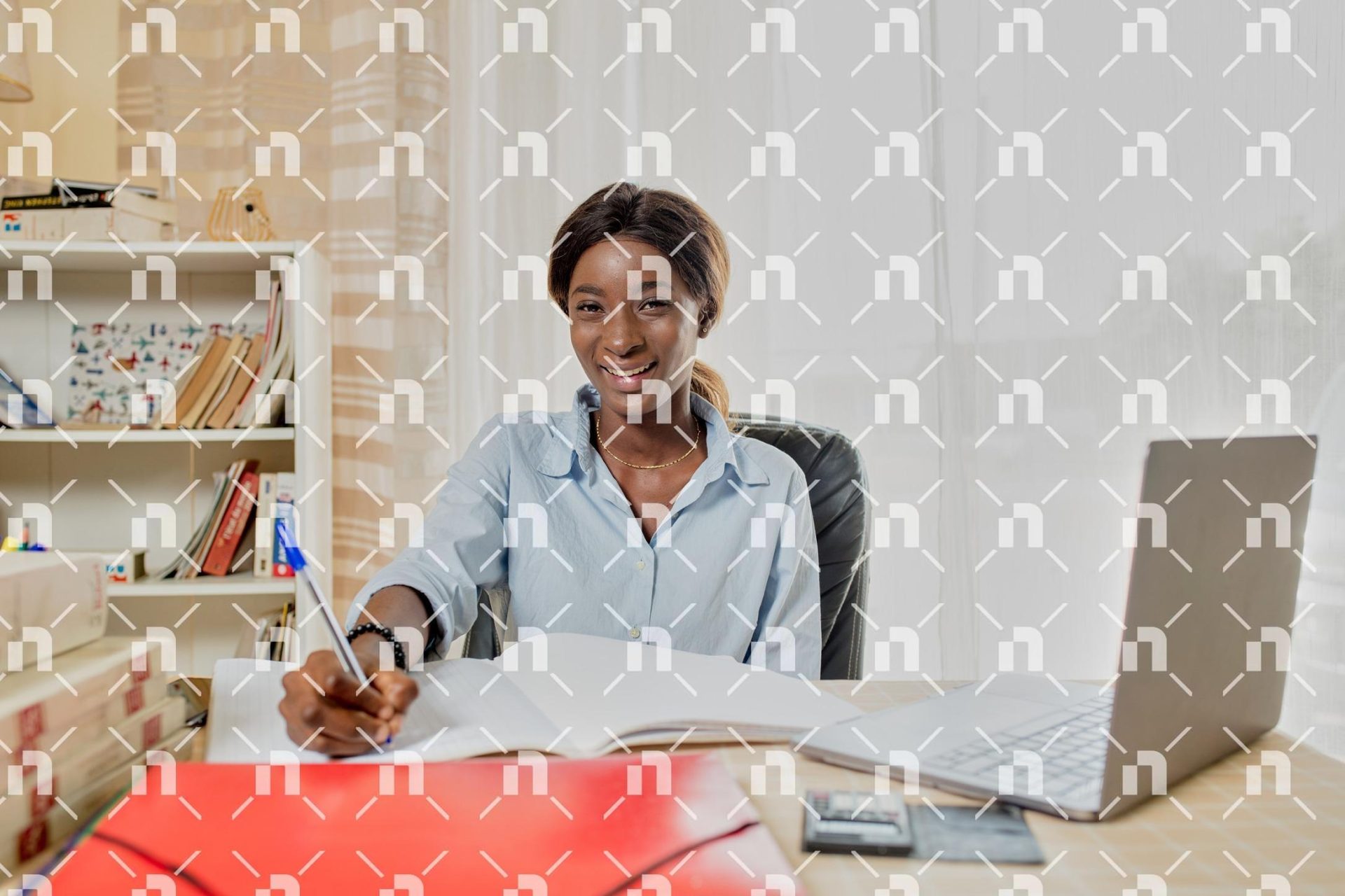 une-jeune-femme-africaine-souriante-assise-a-son-bureau-sur-lequel-est-pose-son-ordinateur-portable-et-ecrivant-dans-un-grand-cahier-avec-un-stylo-bleu